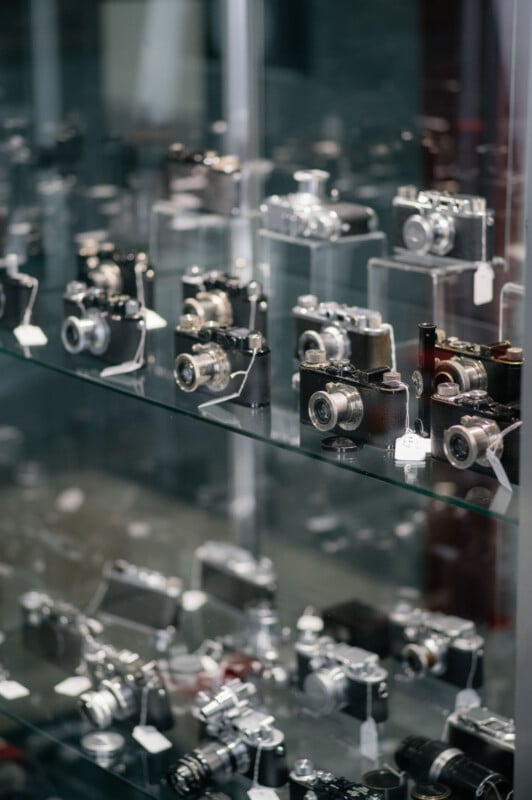 A glass display case filled with multiple rows of vintage cameras, each with small white tags attached, reflecting light and showing various styles and designs.