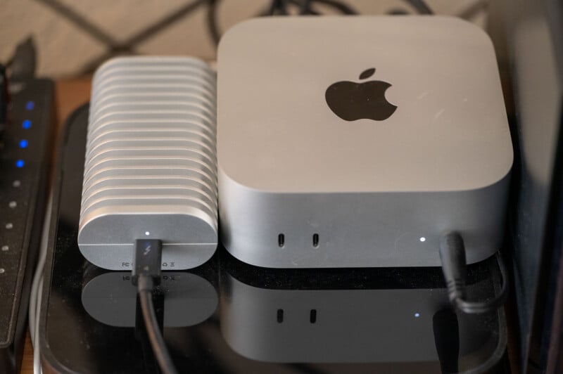 A silver Apple Mac Mini is placed next to a ridged external hard drive on a black surface. Both devices have cables connected and small indicator lights turned on.