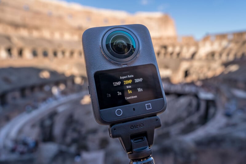 A 360-degree action camera on a tripod is shown close up with its screen displaying photo settings. In the blurred background, the ancient ruins of the Colosseum in Rome are visible.