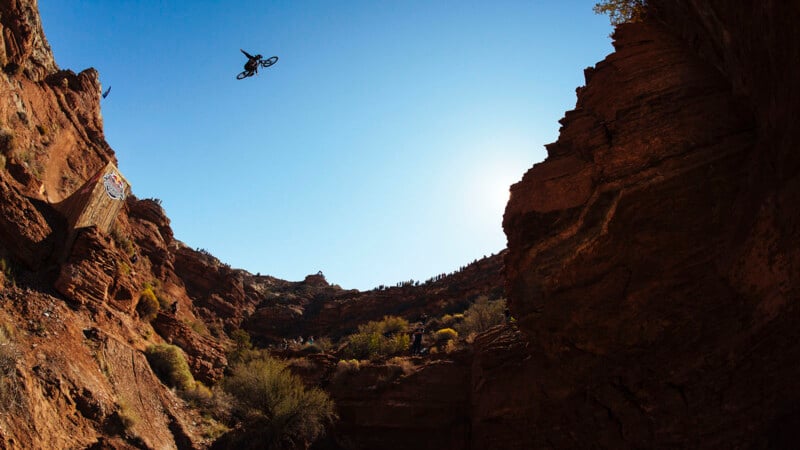 A mountain biker performs a high jump off a ramp built into rugged red rock cliffs, soaring through the clear blue sky above a dramatic desert canyon landscape.