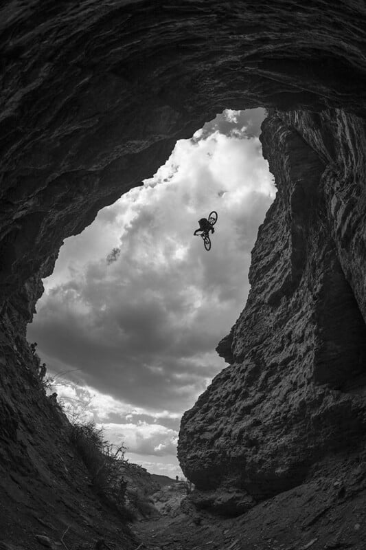A cyclist performs a high jump on a bike between rocky cliffs, viewed from below inside a cave, with dramatic clouds in the sky. The image is in black and white.