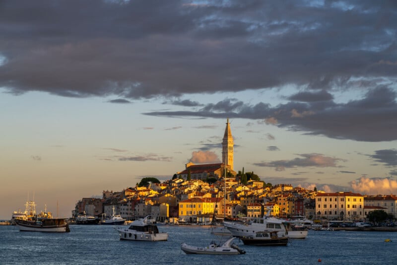 A coastal town with colorful buildings and a tall church tower on a hill, lit by golden sunlight. Several boats float on the calm water in the foreground under a dramatic, cloudy sky.