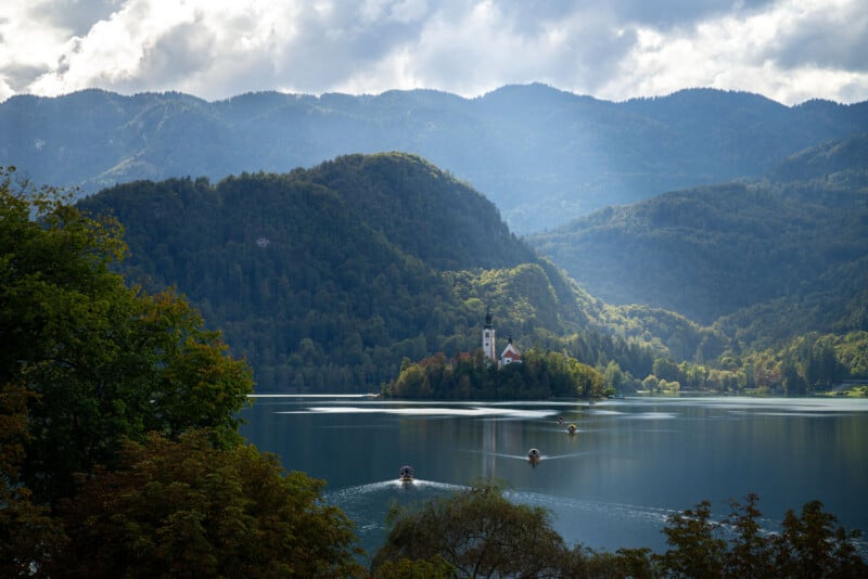 A scenic view of Lake Bled with small boats on the water, an island with a church, and forested mountains in the background under a partly cloudy sky.