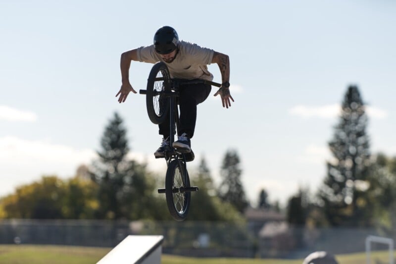 Un hombre con casco realiza acrobacias en una bicicleta BMX en el aire sobre la rampa de un parque de patinaje al aire libre, con árboles y vallas visibles al fondo.