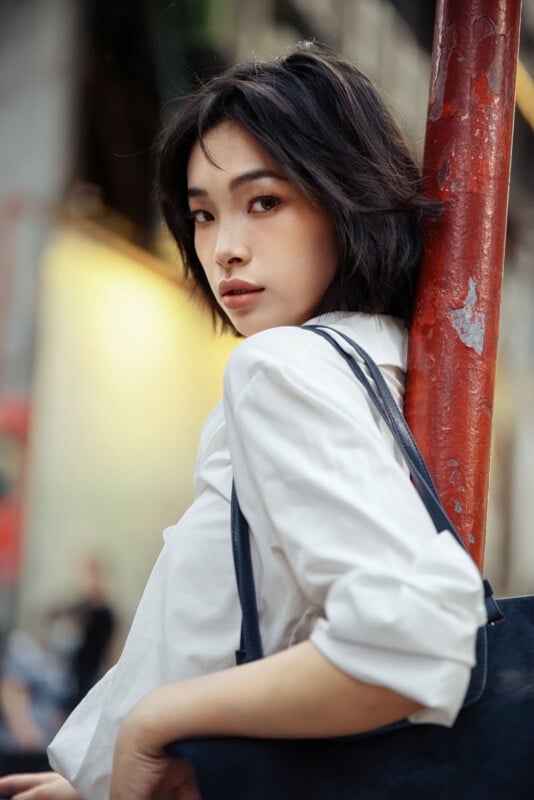 A young woman with short dark hair, wearing a white shirt and carrying a black bag, stands outdoors leaning against a red pole, looking over her shoulder towards the camera.