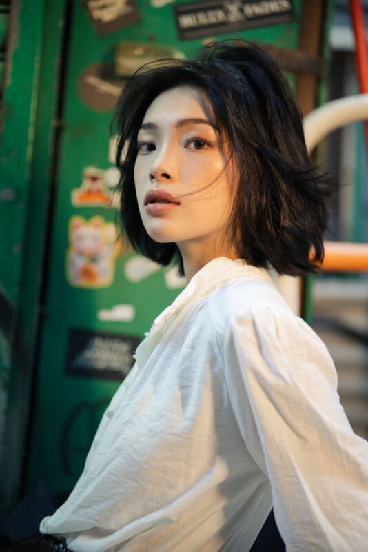 A woman with short black hair and a white blouse poses confidently in front of a green wall decorated with various colorful stickers.