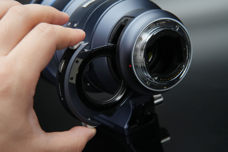 Una mano inserta o retira un filtro o anillo adaptador de la parte posterior de una lente de cámara grande que descansa sobre una superficie negra reflectante.