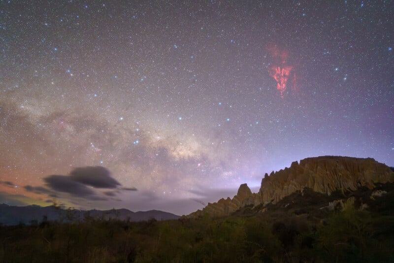 A star-filled night sky with a visible section of the Milky Way above rocky cliffs. Wispy clouds and red streaks of atmospheric light appear, with soft silhouettes of landscapes in the foreground.