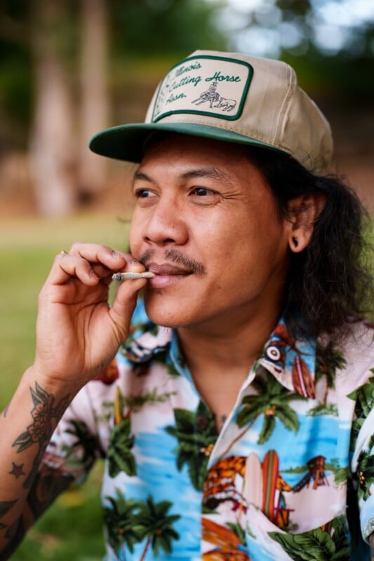 Un hombre vestido con una camisa con estampado tropical y un sombrero beige remendado se sienta al aire libre con un cigarrillo o un cigarrillo de marihuana colgando de su boca. Tiene pelo largo, bigote y tatuajes en los brazos. Las plantas verdes del fondo se vuelven borrosas.