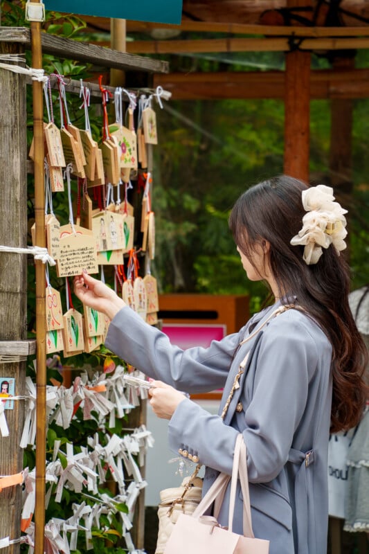 A woman with long dark hair wearing a light blue coat and large hair bow reads wooden prayer plaques (ema) hanging on a display at a Japanese shrine. She holds a paper slip in one hand and stands outdoors by the shrine.