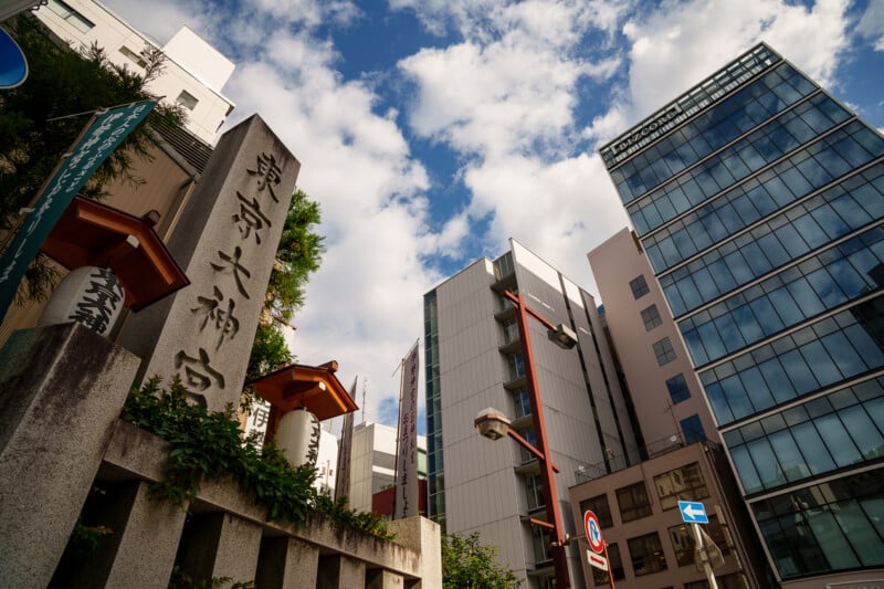 A stone monument with Japanese inscriptions stands beside modern glass and steel buildings under a partly cloudy sky in an urban area.