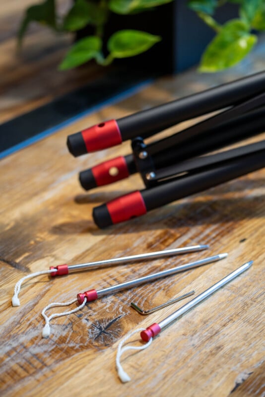 Three black tripod legs with red ends lie on a wooden surface next to three metal stakes with cords and an L-shaped hex key; green leaves are visible in the background.