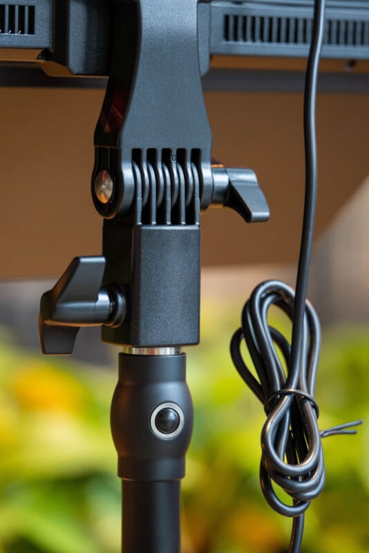 Close-up of an adjustable black metal clamp and joint mechanism holding a device, with a coiled cable hanging on the side; blurred green and yellow background.