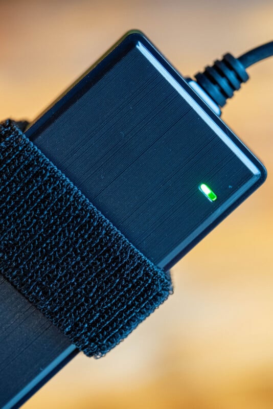 A close-up of a black electronic device with a textured strap around it and a small green indicator light illuminated, connected by a cable in the top corner.