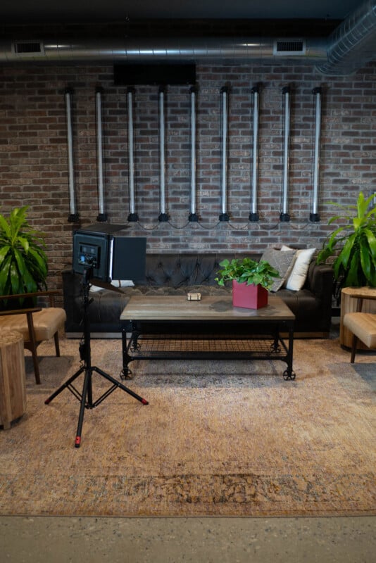 A modern industrial-style lounge with a black tufted sofa, wooden chairs, a coffee table with a plant, and a photography light on a tripod, set against a brick wall with vertical metal pipes.