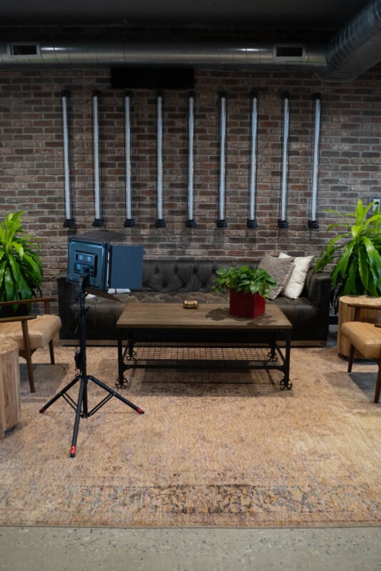 A modern living room with a black leather sofa, a wooden coffee table with a potted plant, tan armchairs, green houseplants, exposed brick wall, vertical metal pipes, and a studio light on a tripod.
