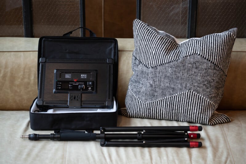 A black LED panel light in an open carrying case, a black tripod with red accents, and a black-and-white patterned pillow are arranged on a beige sofa.