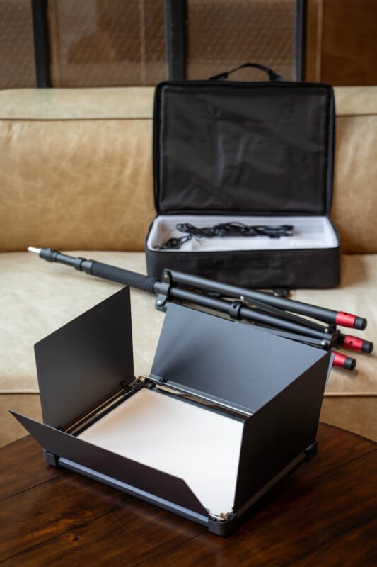 A portable scanning device with an open document holder and a camera mount sits on a wooden table. Behind it, a black carrying case with accessories is open on a beige couch.