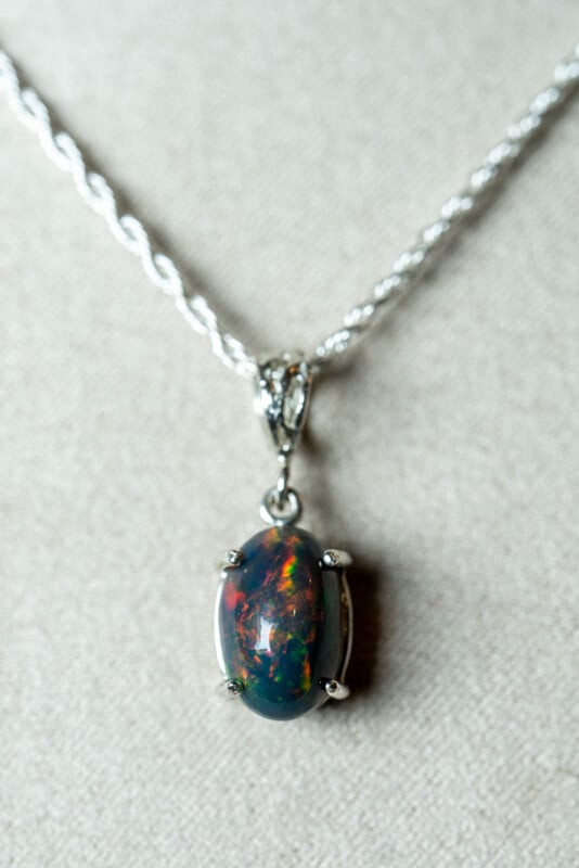 A silver necklace with a twisted chain features an oval-shaped, multicolored opal pendant set in a four-prong silver setting, displayed on a light beige fabric background.