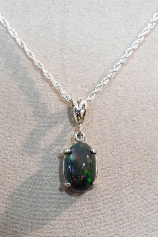 A silver chain necklace with an oval-shaped, dark gemstone pendant displaying green and red iridescent specks, set in a four-prong silver setting, on a light gray background.
