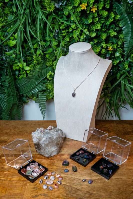 A jewelry display bust with a pendant necklace stands on a wooden table, surrounded by assorted colorful stones, crystals, and three clear display boxes, with a lush green plant wall in the background.