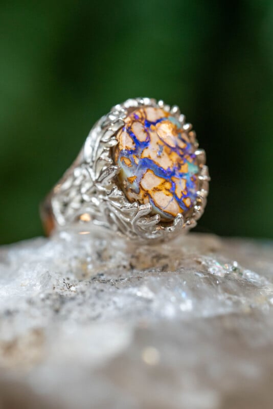 A silver ring with an ornate, textured band features a large, round stone displaying vibrant blue, orange, and brown patterns, resting on a clear, rough crystal surface with a green background.