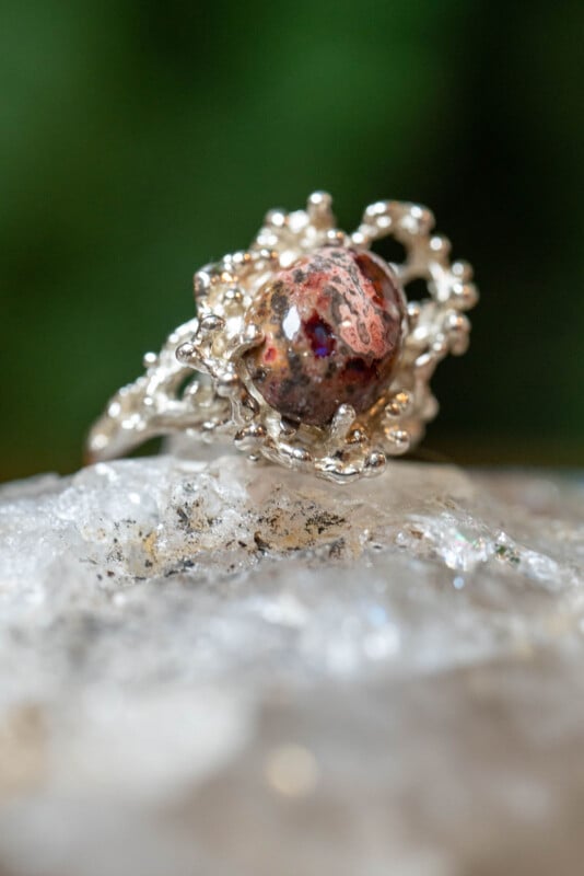 A close-up of a silver ring with an organic, branch-like design, featuring a round, earthy red gemstone, is displayed on a sparkling, translucent surface with a blurred green background.