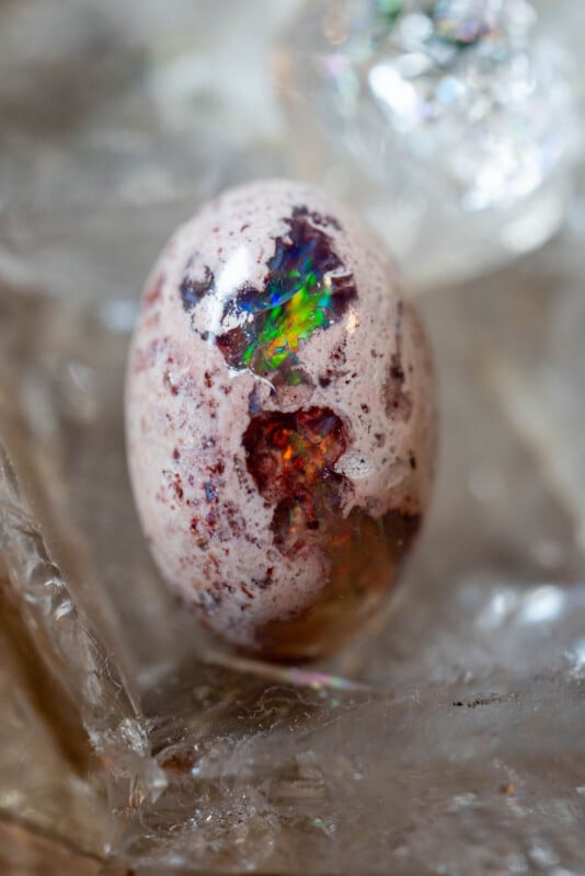 A polished opal gemstone resembling a speckled egg, with vivid flashes of green, red, and blue colors shining through its surface, resting on a textured, translucent background.