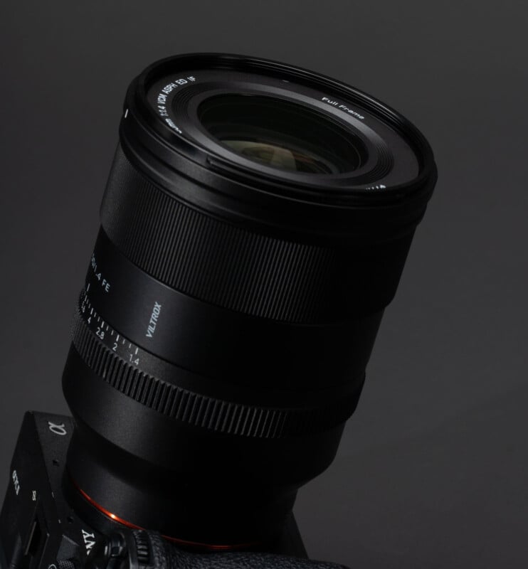 A close-up photo of a black camera lens attached to a camera body, set against a dark gray background. The lens details, such as focal length markings, are visible on the rim.