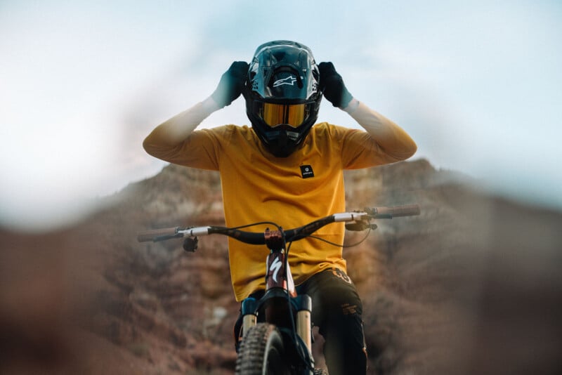 A person in a yellow shirt and black gloves adjusts their helmet while sitting on a mountain bike, with rocky hills and blue sky in the background.