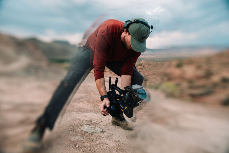 A person in a red shirt, cap, and headphones crouches on a rocky terrain, holding a professional video camera close to the ground, with a blurred effect around the edges of the image.