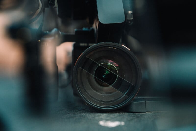 Close-up view of a professional camera lens, focusing on the glass elements and reflections inside the lens. The background is blurred, drawing attention to the circular shape and details of the lens.