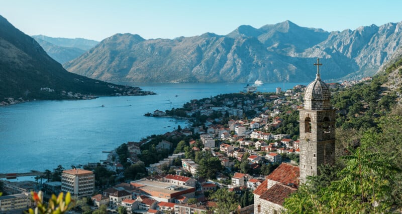 A scenic view of a coastal town with red-roofed buildings beside a bay, surrounded by mountains. A stone church tower with a cross stands in the foreground under a clear blue sky.