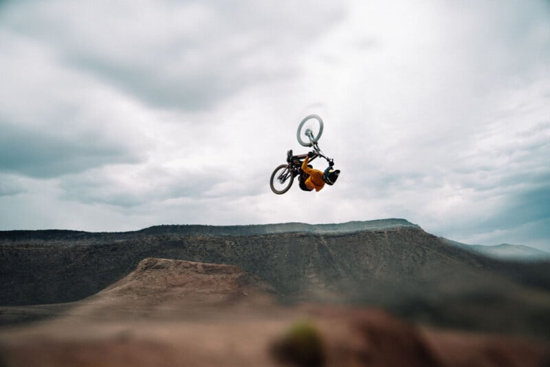 A mountain biker in mid-air performs a backflip over a rocky desert landscape, with distant hills and a cloudy sky in the background.