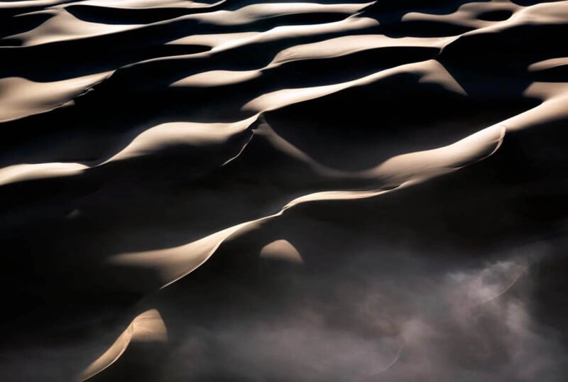 Aerial view of sunlit sand dunes casting soft shadows, creating smooth, flowing patterns and contrasts between light and dark surfaces in a desert landscape.