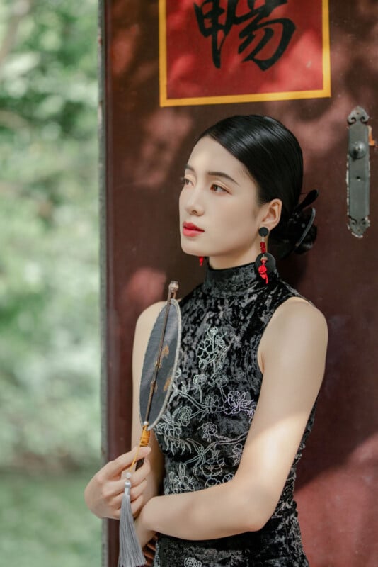 A woman in a black, embroidered cheongsam stands by a wooden door with Chinese calligraphy, holding a round fan. She has sleek, styled hair and wears red earrings, gazing thoughtfully into the distance.