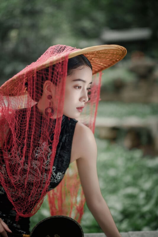 A woman in a traditional black dress and large straw hat draped with a red lace veil poses outdoors amidst greenery, looking pensively to the side.