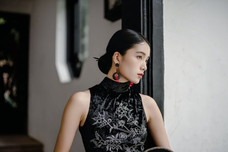 A woman with dark hair in a low bun, wearing a black velvet sleeveless dress with floral embroidery and red earrings, stands by a window looking thoughtfully outside.