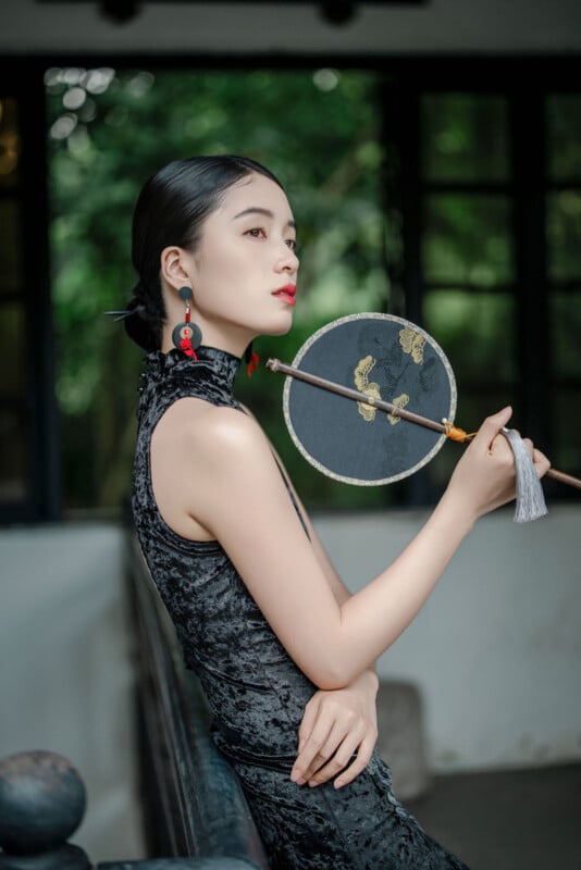 A woman in a sleeveless black velvet dress poses indoors, holding a round embroidered fan near her face. She has sleek, dark hair pulled back and wears red and black earrings, with greenery visible through the window behind her.
