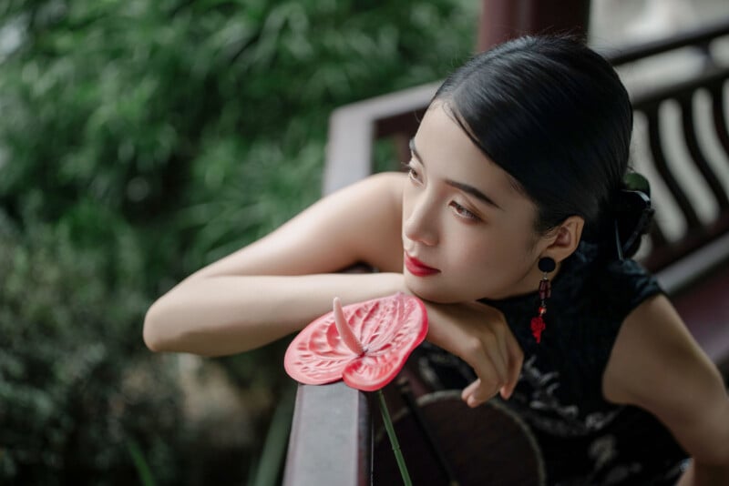 A woman with dark hair leans on a balcony railing, gazing thoughtfully into the distance. She holds a red decorative flower and wears elegant earrings, with greenery blurred in the background.