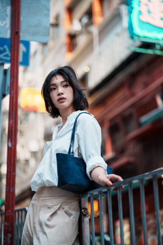 A woman with short dark hair, wearing a white blouse and beige pants, leans casually on a metal railing in an urban street with neon signs and blurred buildings in the background. She carries a navy shoulder bag.