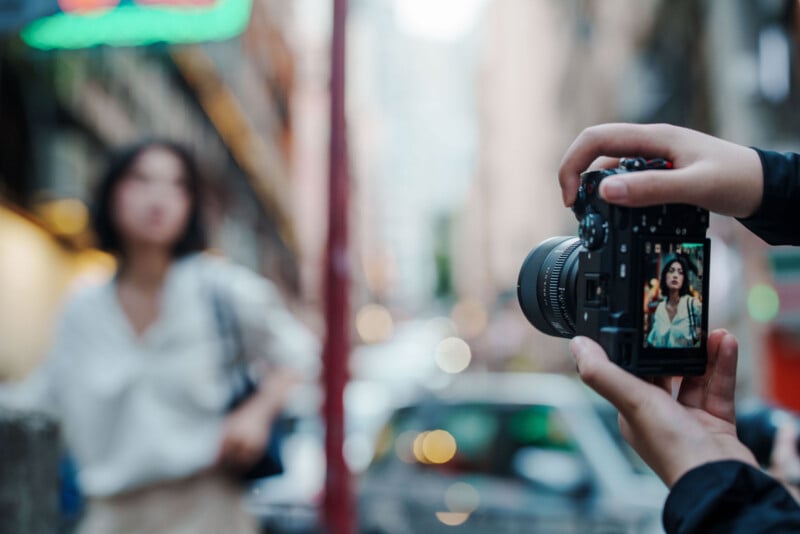A person holds a camera, focusing on a woman standing on a city street. The woman appears sharp on the camera screen but is blurred in the background. Urban buildings and parked cars are visible.