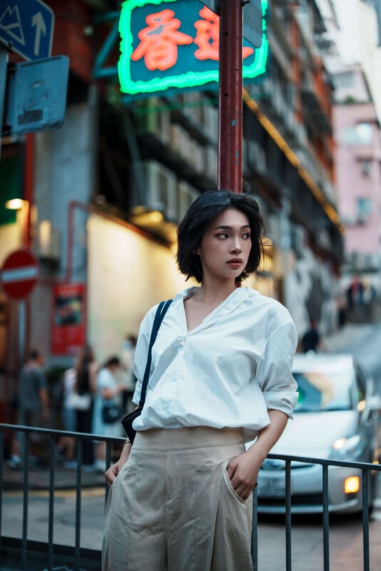 A woman with short dark hair, wearing a white shirt and beige pants, stands by a railing on a city street with neon signs, blurred cars, and people in the background.