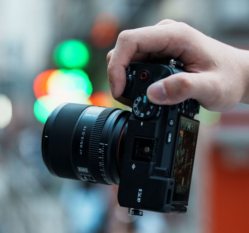 A close-up of a hand holding a black digital camera with a large lens; colorful blurred lights and cityscape are visible in the background.