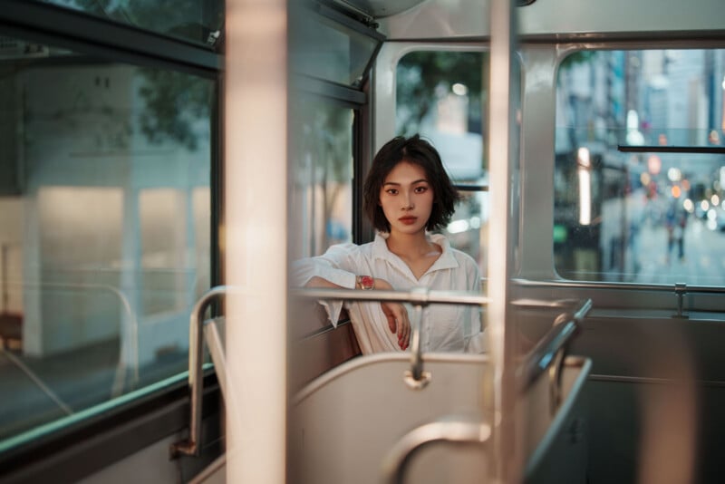 A young woman with short dark hair, wearing a white shirt, sits alone on a bus. The city is visible through the window, and the bus interior is softly lit in the evening. She gazes directly at the camera.