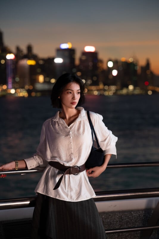 A woman in a white blouse and dark skirt stands by a railing at night, with a city skyline and bright lights blurred in the background across the water.