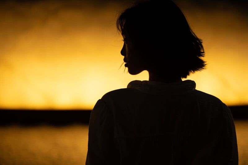 A silhouette of a person with short hair, facing sideways, stands against a glowing yellow-orange background. Their features are mostly in shadow, emphasizing the contrast between figure and light.