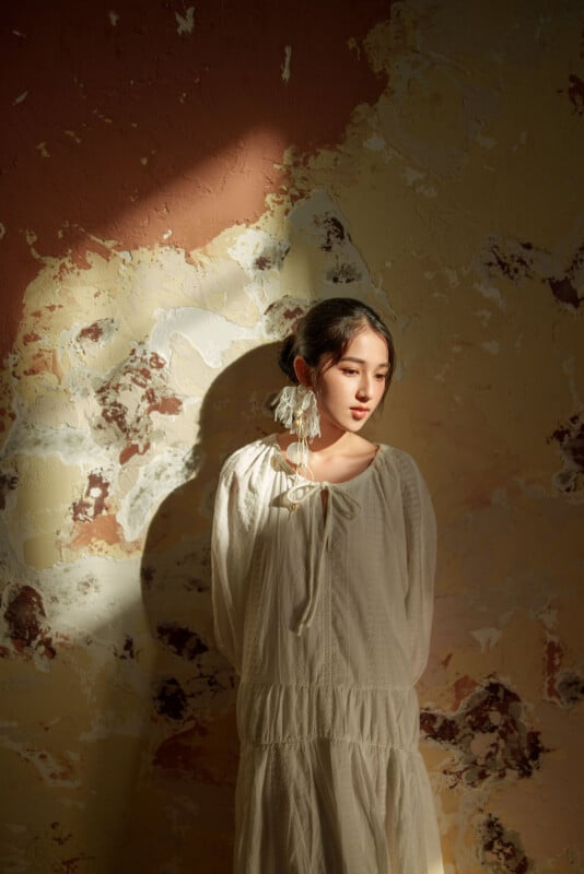 A woman in a long, loose white dress stands against a textured, sunlit wall with peeling paint. She looks to the side, partially in shadow, with large earrings and her dark hair tied back.