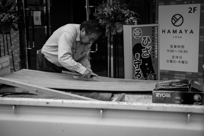 Un hombre con camisa de manga larga cortaba madera con una sierra en la parte trasera de un camión estacionado afuera de la tienda, con carteles japoneses y flores visibles al fondo. La imagen está en blanco y negro.