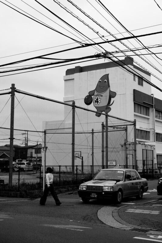 Una escena callejera en blanco y negro muestra a un hombre cruzando la calle cerca de un taxi con cables sobre su cabeza. Una gran mascota de pez de dibujos animados sosteniendo una pelota de baloncesto está pintada en el edificio del fondo.